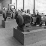 Maschinenraum eines Wasserwerks mit mehreren elektrischen Kreiselpumpen auf Betonsockeln und einem Bediener an der Schalttafel.