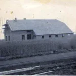 Schwarzweißfoto eines im Bau befindlichen Wohnhauses mit Gerüst in der Siedlung Walchumer Moor, umgeben von Feldern.