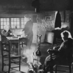 Schwarzweißfoto: Drei Personen in einer ärmlichen Steinhütte um ein offenes Feuer am Boden, 1950er Jahre.
