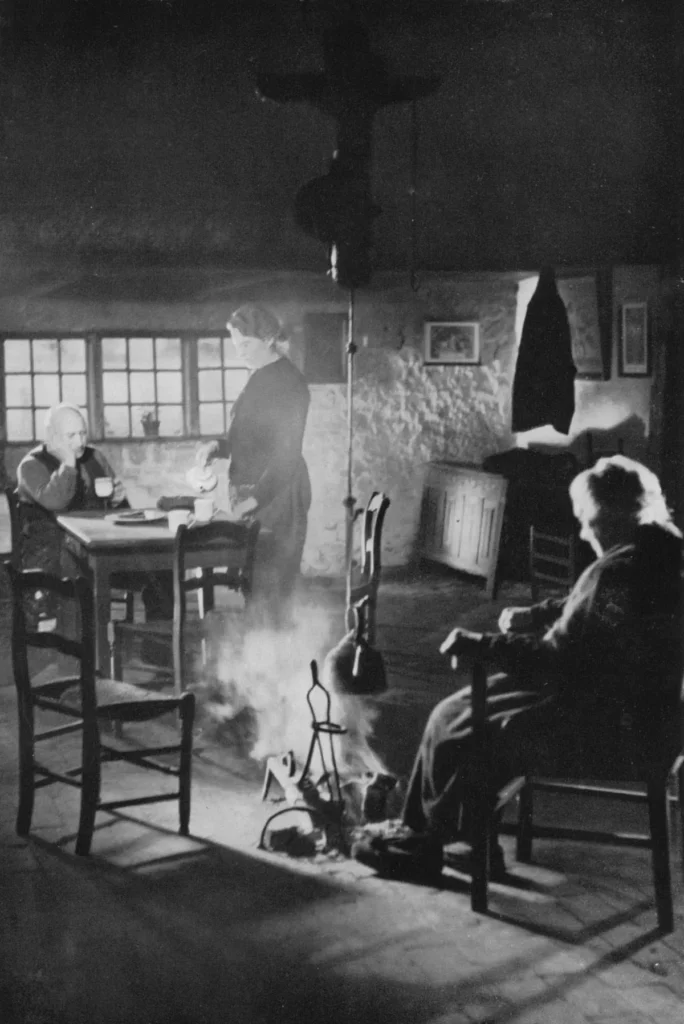 Schwarzweißfoto: Drei Personen in einer ärmlichen Steinhütte um ein offenes Feuer am Boden, 1950er Jahre.