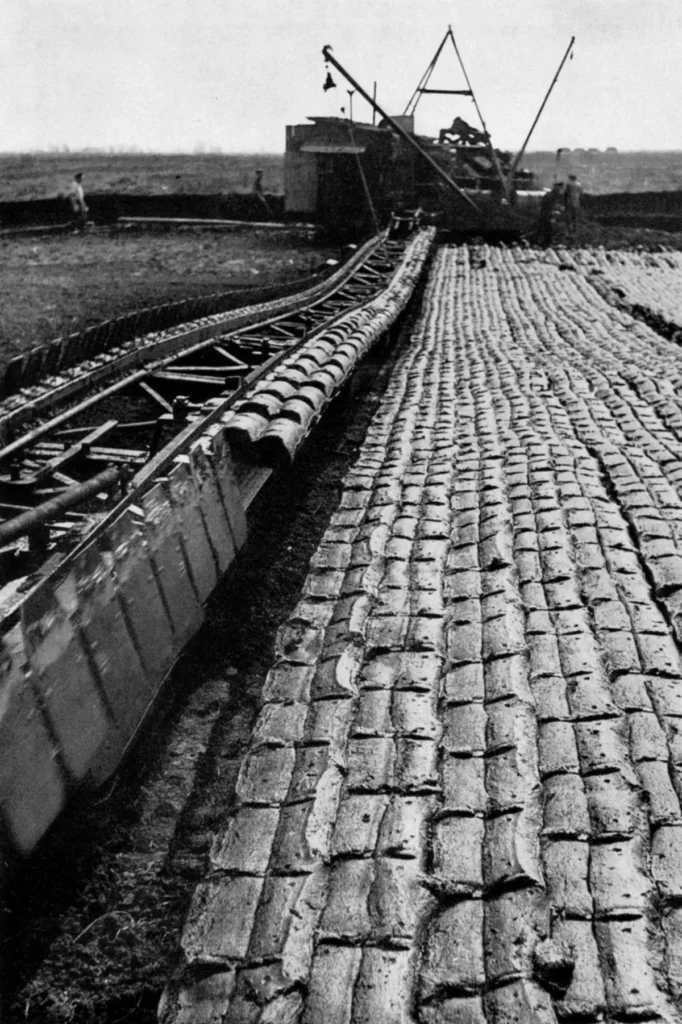 Historische Schwarzweißaufnahme von maschinellem Torfabbau im Emsland mit Förderbahn und gestapelten Torfziegeln.